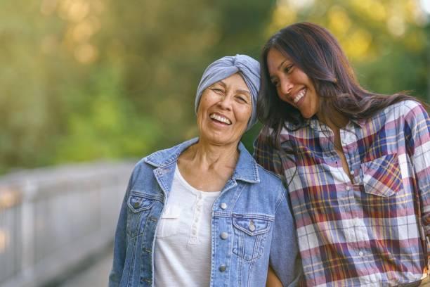 An woman embraces her senior mother with cancer. They are outside and both are smiling and happy to be together. The daughter is looking toward her mom affectionately.