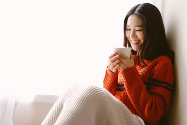 Portrait of beautiful Asian woman relaxing with cup of hot cocoa or chocolate with marshmallow while sitting by the window. Happy girl enjoying warm beverage in winter day.  Lazy day off concept.