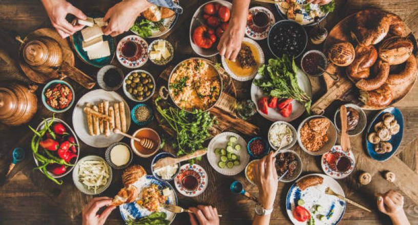 Turkish breakfast table. Flat-lay of peoples hands taking pastries, vegetables, greens, olives, cheeses, fried eggs, spices, jams, honey, tea in copper pot and tulip glasses, wide composition